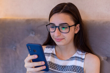 Young Caucasian girl with glasses sitting rest on sofa and browsing internet on smartphone. Stay at home concept