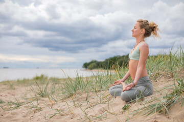 Young blonde woman practicing yoga on the coast. Professional trainer meditating in beach dune by...