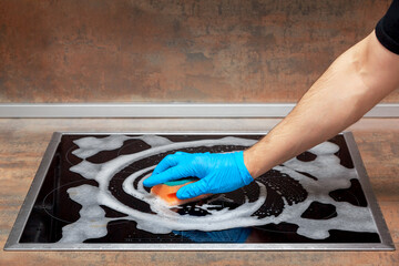 Men's hand in blue glove clean kitchen electric stove with a sponge and cleansing foam