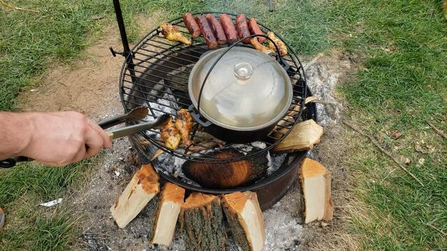 High angle view of sausage and chicken drumstick being cooked on fire pit or campfire alongside cast iron pot of boiling fish soup.