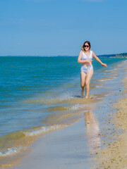 Fototapeta premium A young girl runs along the sandy sea beach along the surf. Splashes of sea water. The girl is wearing a white swimsuit and sunglasses. Freedom and carelessness. Outdoor fitness. Sunny day. Copy space