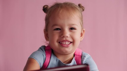 childhood, school, education, upbringing, science concept - close-up happy little blonde caucasian slavic girl in blue dress with backpack hugs book in hands smiling broadly on solid pink background