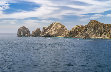 Cabo San Lucas, Mexico - April 22, 2008: The south end of Baha California is a collection of beige rocks in a gray ocean under blue cloudscape. Tiny beach and small pleasure vessels in front.