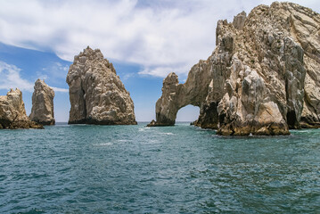 Fototapeta premium Cabo San Lucas, Mexico - April 22, 2008: South end of Baha California. The Arch in beige rocks formation and more free-standing peaks. Greenish ocean water in front under blue cloudscape.