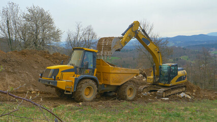 Big excavator unloads a bucket full of soil in the back of a yellow truck.