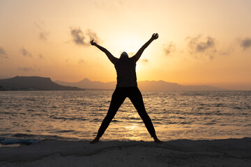 Fototapeta premium Silhouette, young woman with protective surgical face mask performs yoga stretching exercises at the beach at dusk during covid-19 coronavirus pandemic