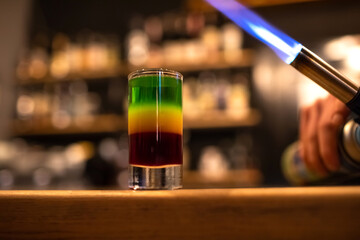 Beautiful tricolor cocktail. Red green yellow alcoholic drink standing on the bar counter, bartender with a lighter prepares to set fire to the glass