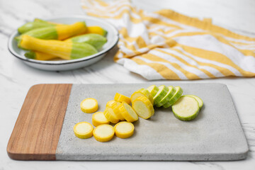 Sliced Zephyr Squash on a Cement Cutting Board on a White Countertop; White Bowl of Whole Zephyr Squash in Background