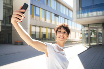 Smiling mature woman taking selfie using smartphone against buildings in city on sunny day