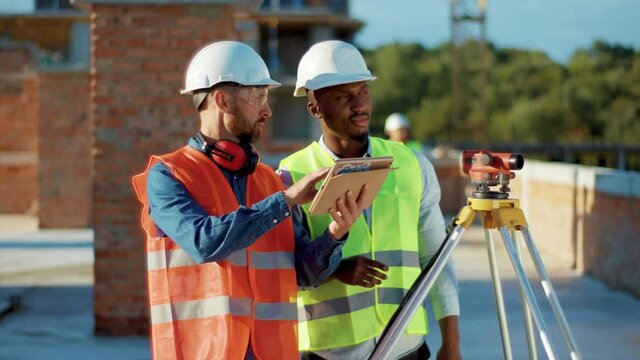 At sunlight two men engineers surveyor take measures with theodolite use tablet computer talking stand helmet hardhat geodesy building engineering real estate teamwork infrastructure industry slow