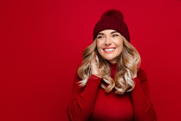 Charming smiling blonde woman touches curly hair with both hands, wears red wool turtleneck and hat, poses against red studio wall, enjoys the winter weather, looking at blank space for advertising