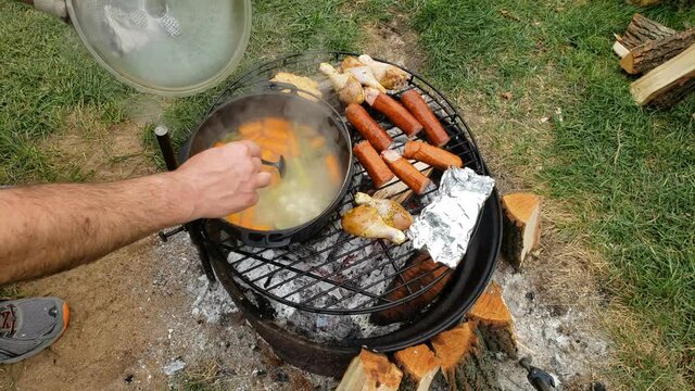 Opening lid of cast iron pot to check and stir soup broth being cooked over fire pit alongside other food like sausage and chicken drumsticks.