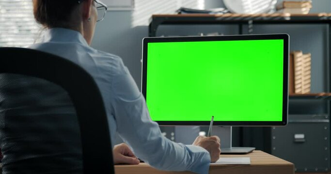 Back view of business woman taking notes during video conference on modern computer with chroma key screen. Online meeting and conversation. Concept of technology.
