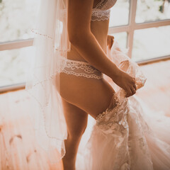 European skinny bride in lingerie puts on a wedding dress. The girl with the veil stuck one leg in the dress.