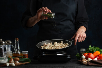 The chef i black uniform adding some spices to the frying pan with chicken isolated on dark background. Cooking tasty dish for dinner. Chef's hands preparing the meal. Food concept.