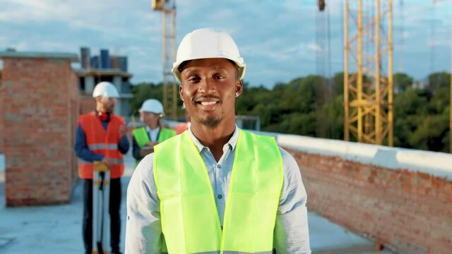 African american young man builder in hard hat standing look at camera smiling on background of building under construction helmet industrial industry structure architecture inspector slow motion