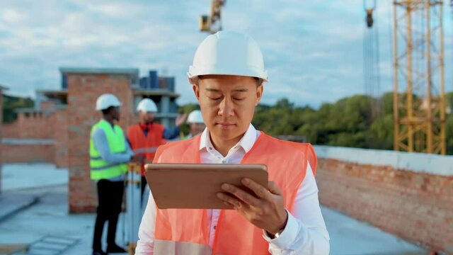 Serious asian builder standing with tablet computer look at camera on background of building under construction helmet industrial portrait industry structure architecture inspector slow motion