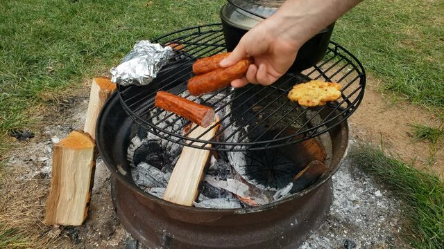 Cooking food like sausage and chicken legs alongside cast iron pot of boiling fish soup using fire pit as heat source.