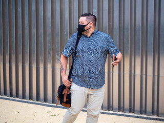 Hombre de negocios joven y tatuado andando por la calle usando su teléfono movil vistiendo mascarilla respetando las medidas de seguridad del coronavirus 