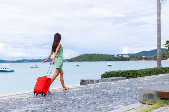 Young happy beautiful woman in dress with red suitcase on vacation by tropical beach 