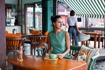 Cute elegant calm happy woman in green summer dress sits with coffee in cafe enjoying morning. Beautiful coffee lover. 