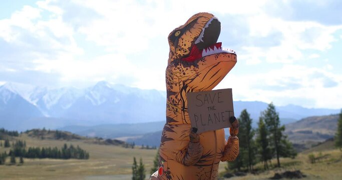 Huge dinosaur doll Tyrannosaurus Rex with person inside is holding banner save the planet in paws. Nature park in mountains. Concept of environmental protection. Installation with prehistoric animal.