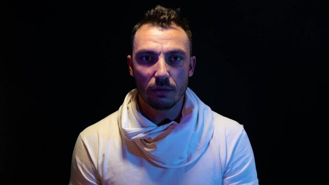 A close-up man in a white t-shirt is in a dark room. Warm light falls from above, creating harsh shadows. The person stands motionless and talks. On a black background.