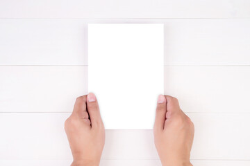 Hand of young man holding poster or card or brochure with white paper blank on wooden table, greeting card, a5, a4, flyer advertising and message for design empty, copy space, mockup template.