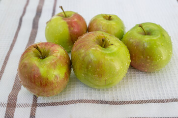 A small pile of freshly washed green-red apples, with water droplets on the surface of the peel, lying picturesquely on a white waffle fabric tablecloth with a border framed by brown stripes.