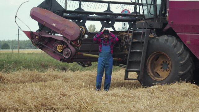Young promising farmer near a combine harvester in a field with cut stalks of grain crops during the harvesting campaign. A professional of his business poses near an agricultural machine