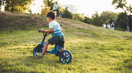 Caucasian boy with a bike is riding on the grass during a walk in park with his parents