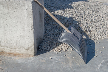 A wide shovel with a wooden handle and a black plastic working part leaning against a concrete block on a flat surface near a pile of gray gravel.