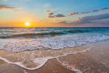 Sea sunset with a sun disc in the sky with clouds and white foam waves on the sand in the foreground.