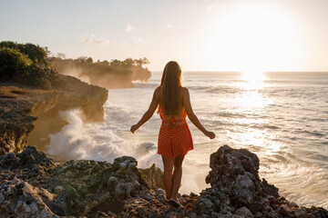 Back view woman standing on a high cliff with sea view, look at sunset and occean wawes crash the rocks