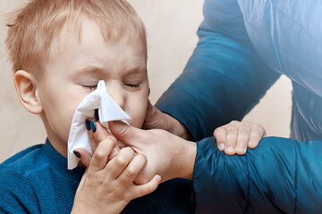 Little blond boy sneezing or blowing his nose with mother helping hands. Squinted eyes, frowning,...
