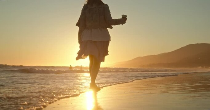 Woman at beach, feeling happy and relaxed. Perfect commercial shot of summer vacation. Slow motion of model walking by the sea waves, drinking cup of coffee and enjoying the view of golden sunset