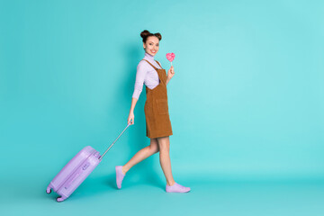 Full length profile photo of attractive two buns hairdo lady tourist check-in terminal arm hold luggage lollipop wear brown mini dress violet shoes pullover isolated turquoise color background