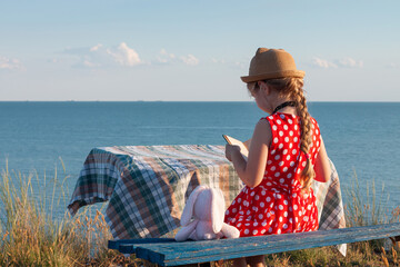 Child girl in hat and polka-dot dress sitting on vintage bench reading a book. Kid looking at Holy Bible in hands and praying on sea lanscape background. Friendship peace religion faith hope concept.