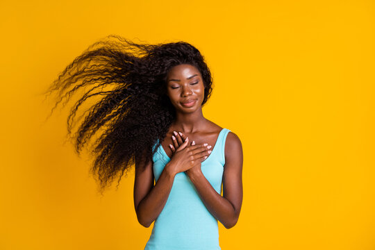 Photo portrait of young curly brunette african american woman holding two hands on chest with closed eyes wearing blue dress isolated on vivid yellow colored background
