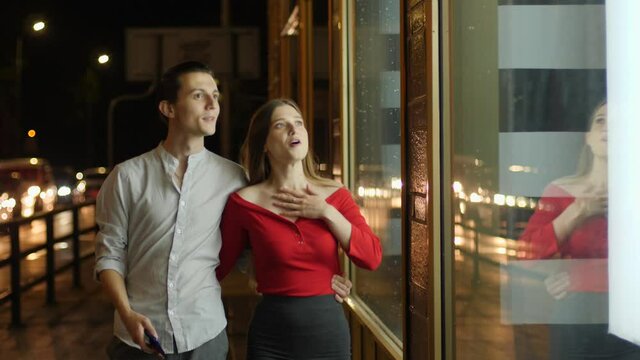 Young happy couple watch store showcase and see expensive thing they wish to buy behing glass frontage during night city walk and take phone picture of it to remember