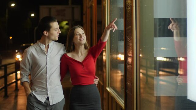 Young happy couple watch store showcase and see expensive thing they wish to buy behing glass frontage during night city walk