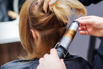 Fototapeta premium Professional hairdresser twists curls of long light brown hair of woman with curling iron in beauty salon. Hairdressing procedures
