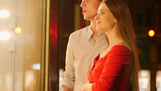 Young happy couple watch store showcase and see expensive thing they wish to buy behing glass frontage during night city walk