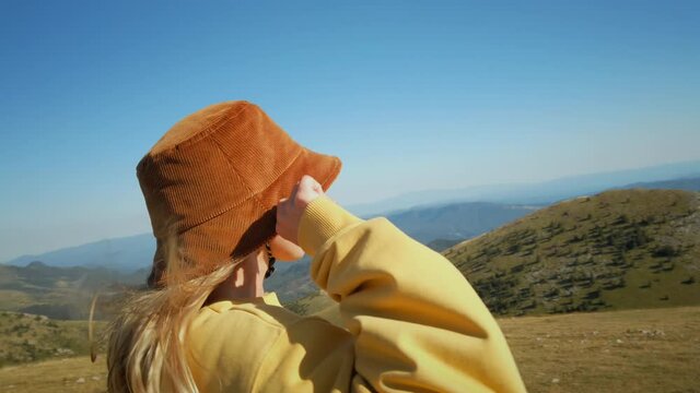 Portrait of laughing and happy young woman in bucket hat stand on top of mountain, bathing and enjoying sun and fresh wind breeze. Smiling and content about spending time in nature