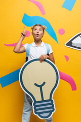 Pensive schoolgirl having idea while holding decorative light bulb near paper art on yellow background