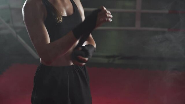 Female fighter wraps her hands with boxing bandages, kickboxing training day in a gym, neon lighting.