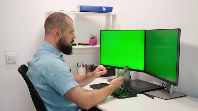 Man Sitting At Home And Texting On Smart Phone Against Two Green Screens Or Monitors. Fingers Typing On Touch Screen