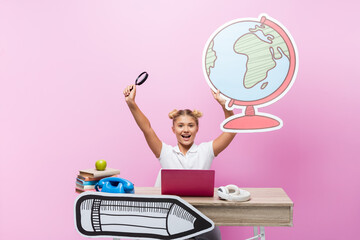 Excited schoolkid holding loupe and paper globe near laptop, headphones and books on pink background