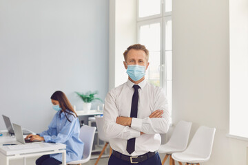 Young company worker wearing face mask standing in his workplace looking at camera