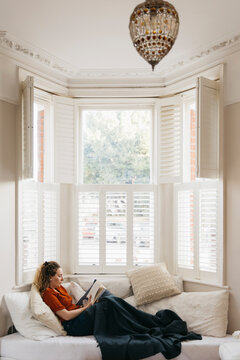 Young woman relaxing on living room sofa reading book in bay window
